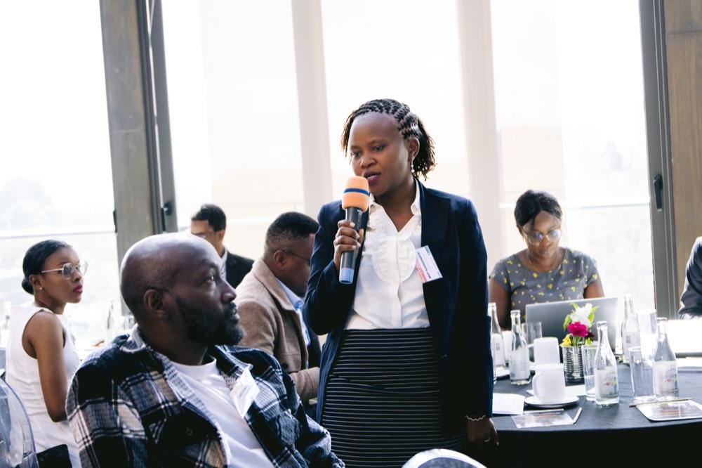 A woman stands to speak into a microphone at a round conference table.