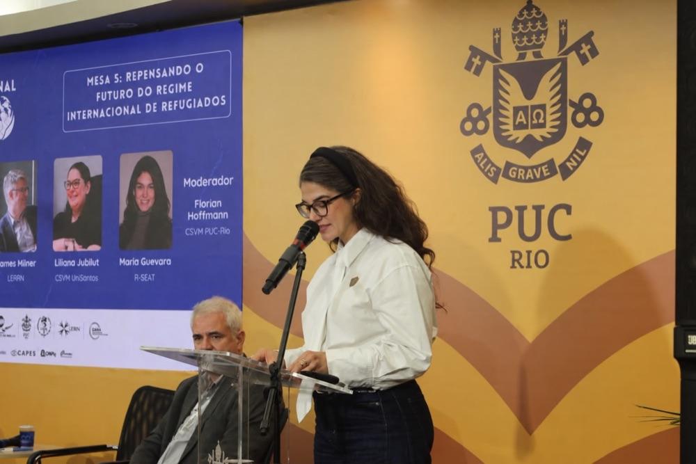 A woman speaks at a lectern on a panel at PUC-Rio.
